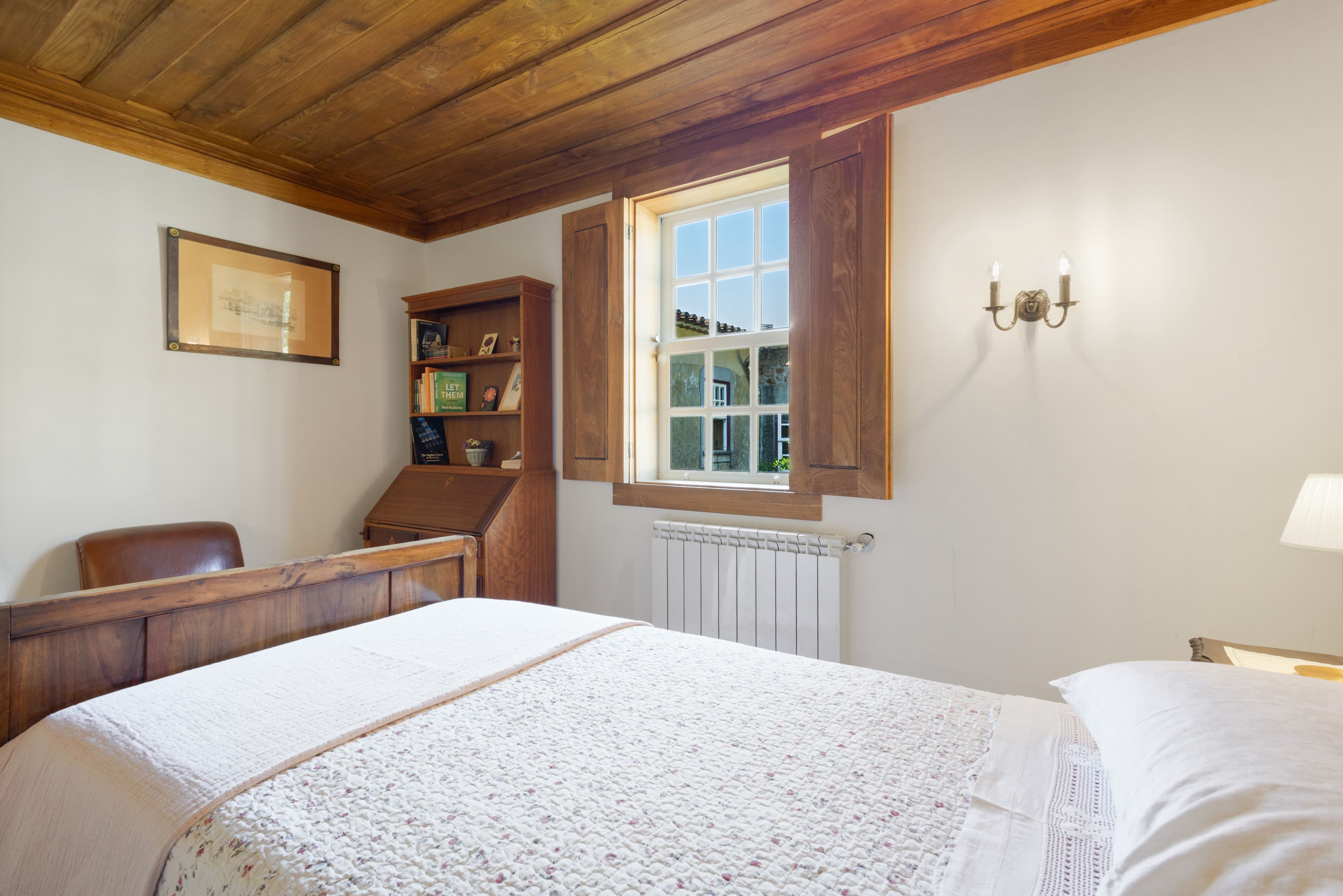 Una camera da letto con soffitto in legno, un letto con copripiumino bianco, una scrivania in legno, una libreria e una finestra con persiana aperta.