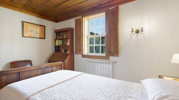 Una camera da letto con soffitto in legno, un letto con copripiumino bianco, una scrivania in legno, una libreria e una finestra con persiana aperta.