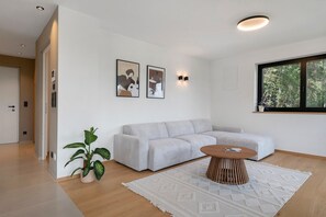 Ein modernes Wohnzimmer mit einer Eckcouch, einem Couchtisch und einem großen Fenster mit Blick auf Grünflächen.