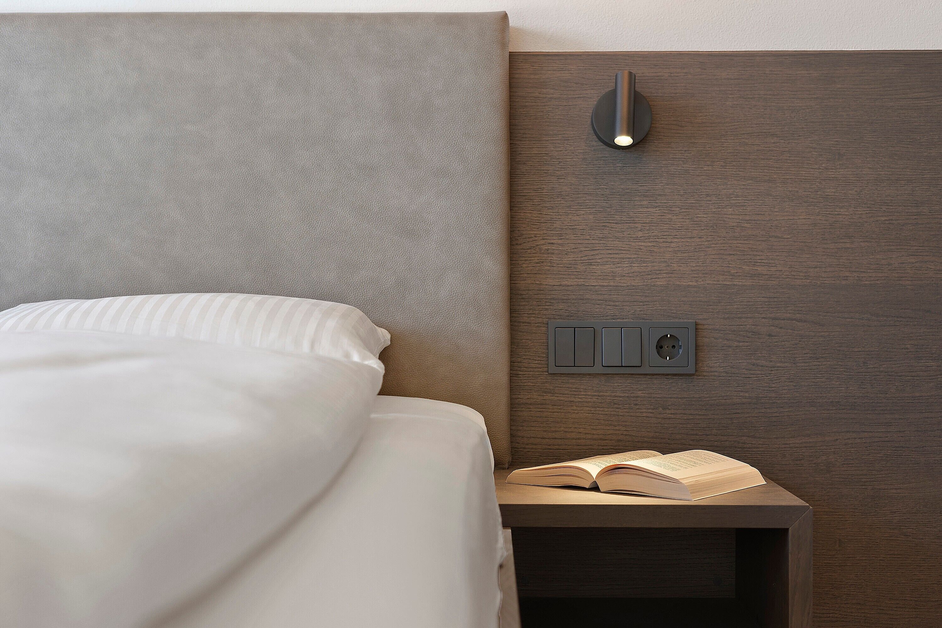 A neatly made bed with a white comforter and pillows, a bedside table with an open book, and a wall-mounted light fixture.