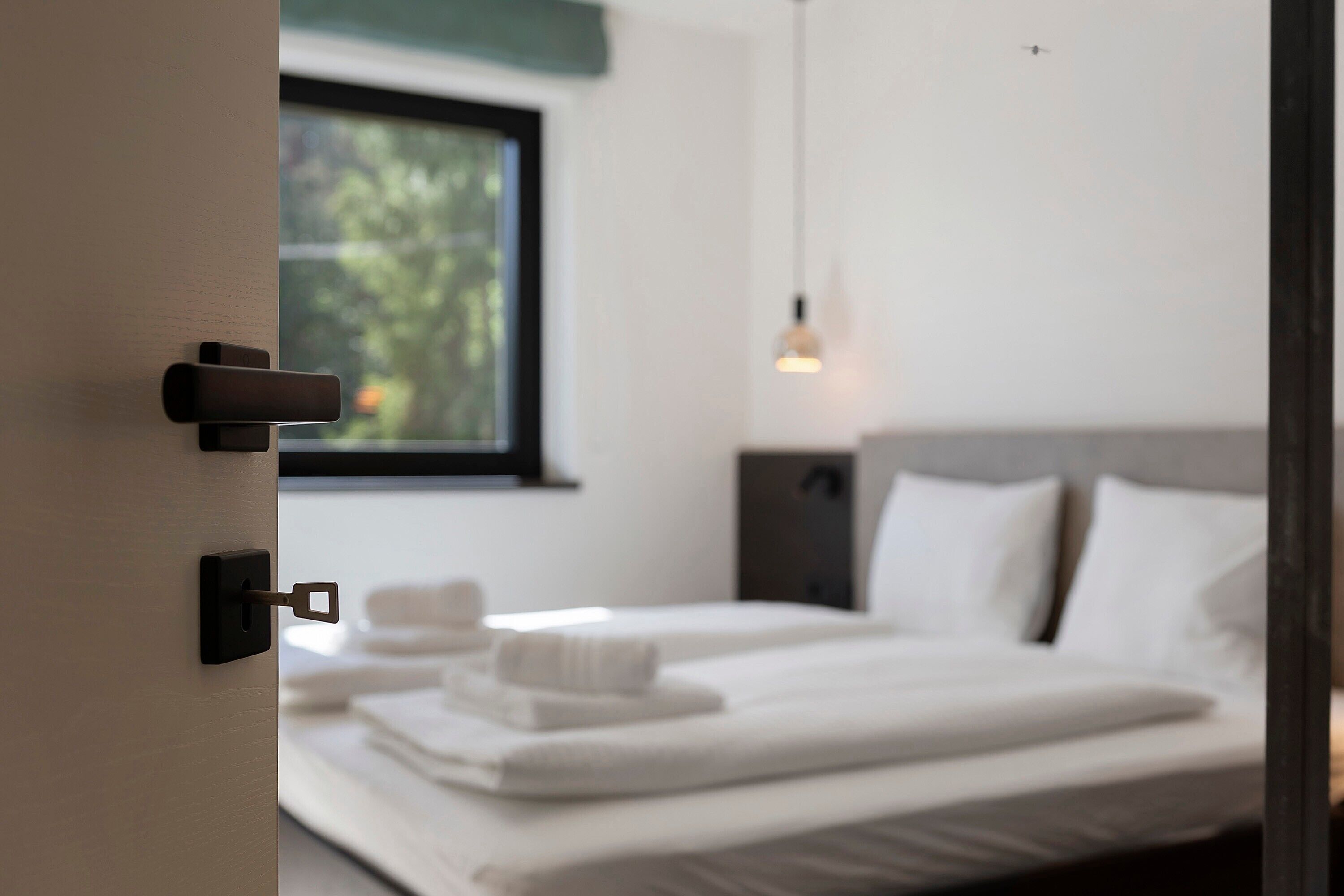 Ein Hotelzimmer mit Bett, Fenster, einer Pendelleuchte und einer Tür mit Schlüsselloch.