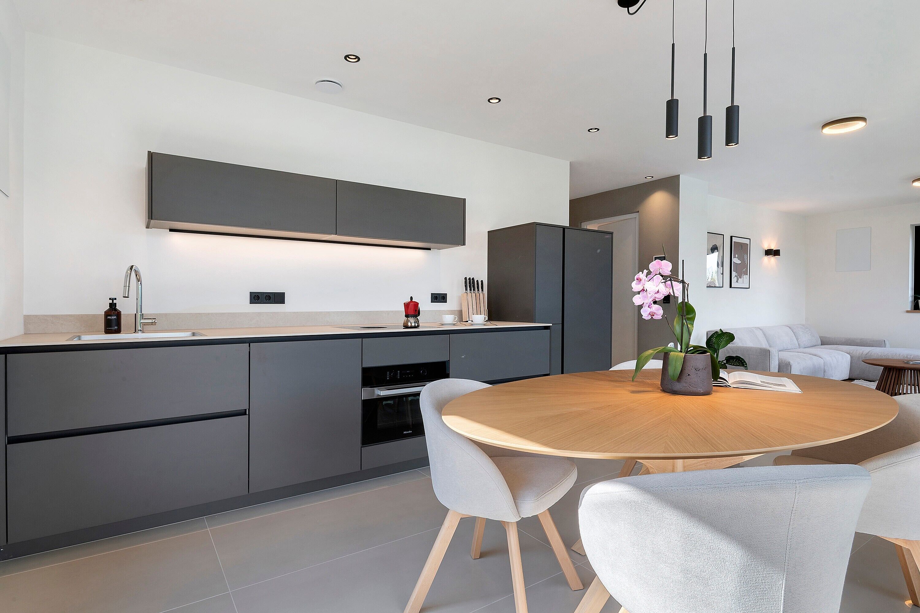 A modern kitchen with grey cabinetry, a built-in oven, and a round wooden dining table with chairs.