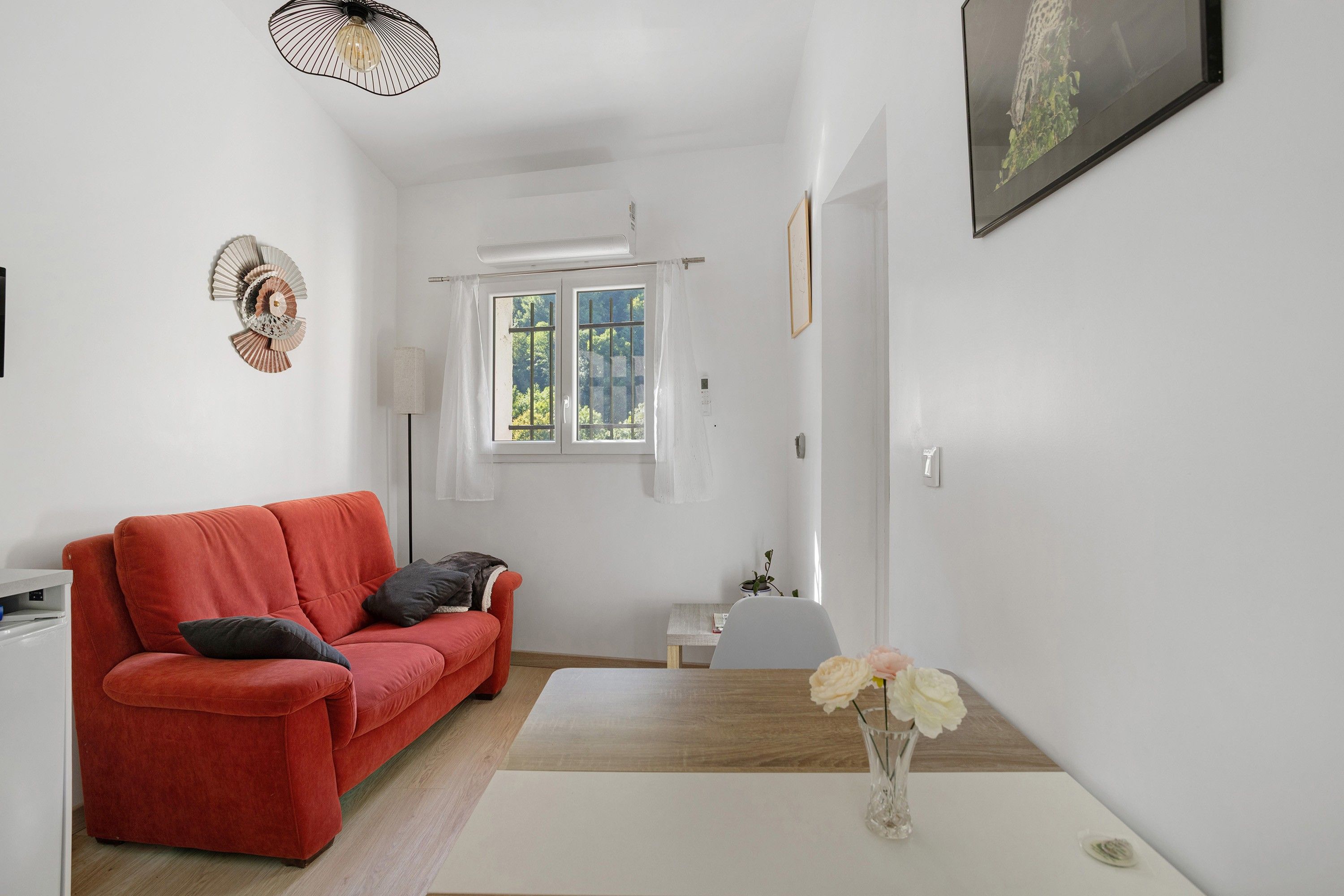 Een kamer met een rode bank, een witte tafel met een bloevas, een raam met uitzicht op groen en een aan de muur hangend schilderij.