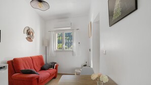 A room with a red sofa, a white table with a vase of flowers, a window with a view of greenery, and a wall-mounted picture.