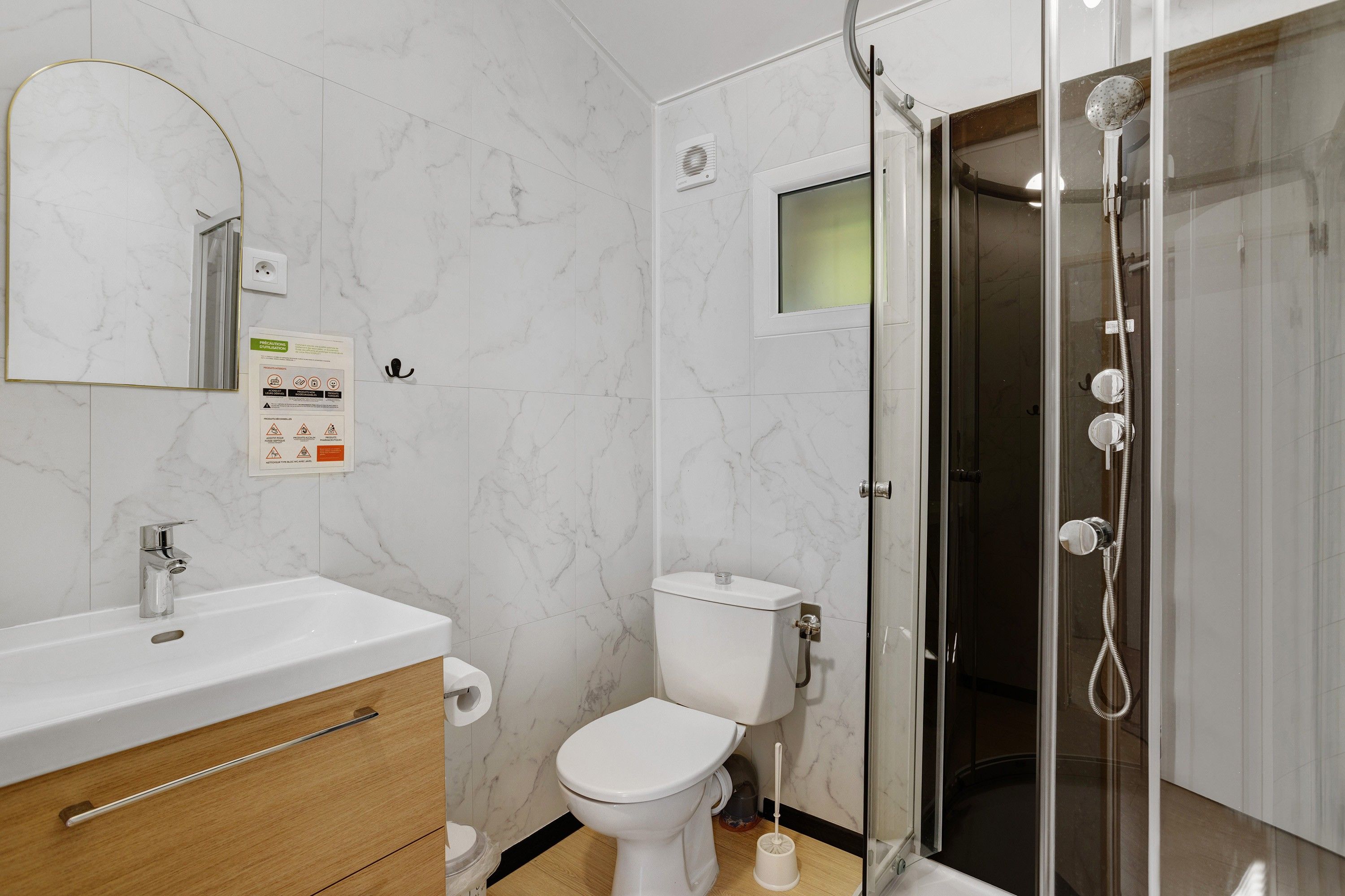Un baño con pared de mármol blanco, cabina de ducha de vidrio, inodoro blanco y tocador de madera con espejo.