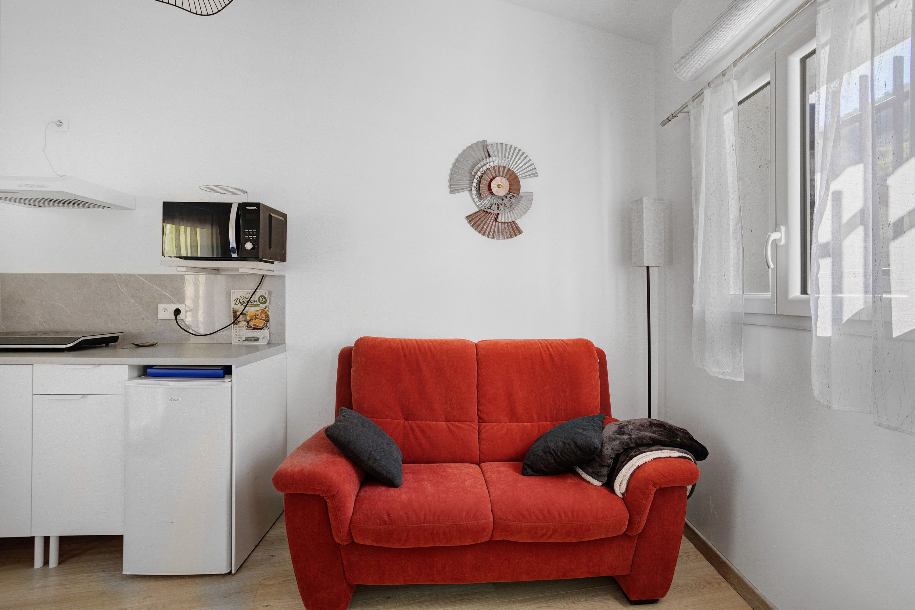 Een ruimtelijk ingerichte leefruimte met een kitchenette, een rode bank met kussens en een decoratief wandobject.
