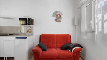 Een ruimtelijk ingerichte leefruimte met een kitchenette, een rode bank met kussens en een decoratief wandobject.