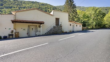 Ein Gebäude mit einer Steintreppe, die zu einer Tür führt, einer gepflasterten Straße davor und einem bewaldeten Hang im Hintergrund.