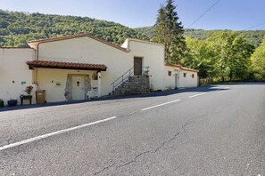 A building with a stone staircase leading to a door, a paved road in front, and a forested hillside in the background.
