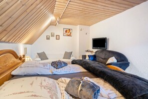 Ein gemütlicher Raum mit schrägverdeckter Decke, einem Bett mit floralem Bettzeug, einem dunklen Sofa und einem flachbildfernseher an der Wand.