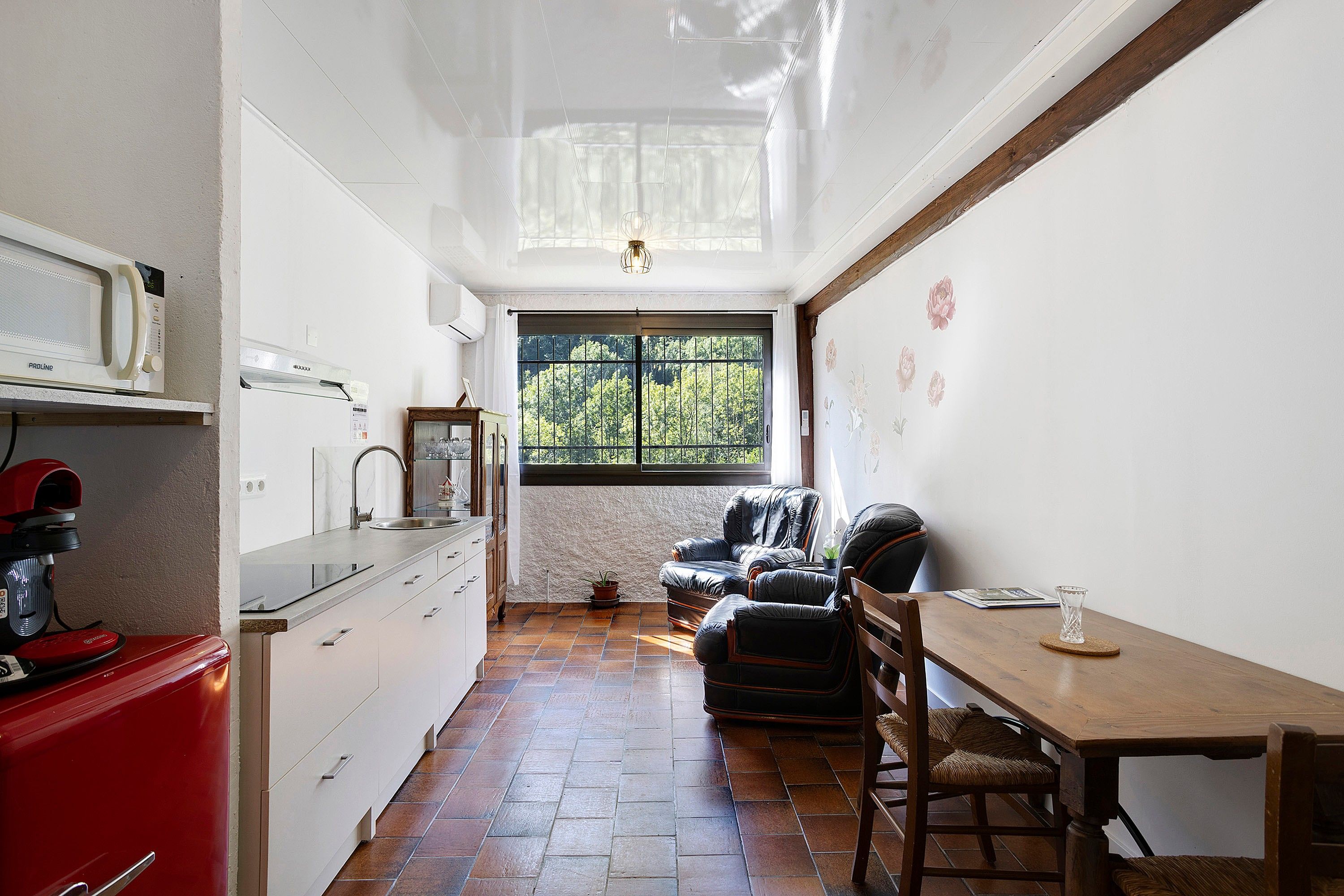 Een smalle keuken met een raam, een eethoek met een tafel en stoelen, en een rode apparatuur aan de linkerkant.