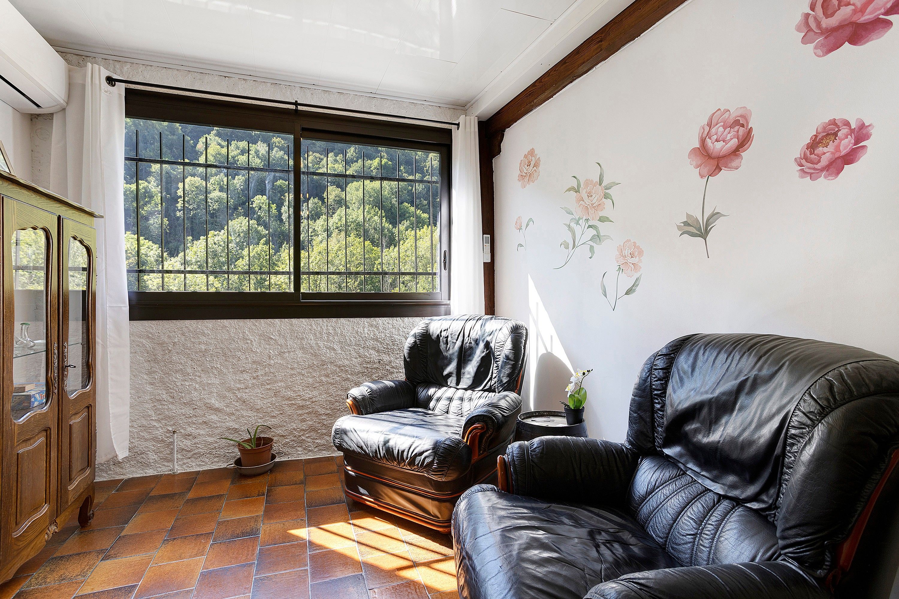 A room with a large window, a leather armchair, a wooden cabinet, and a floral wall decoration.