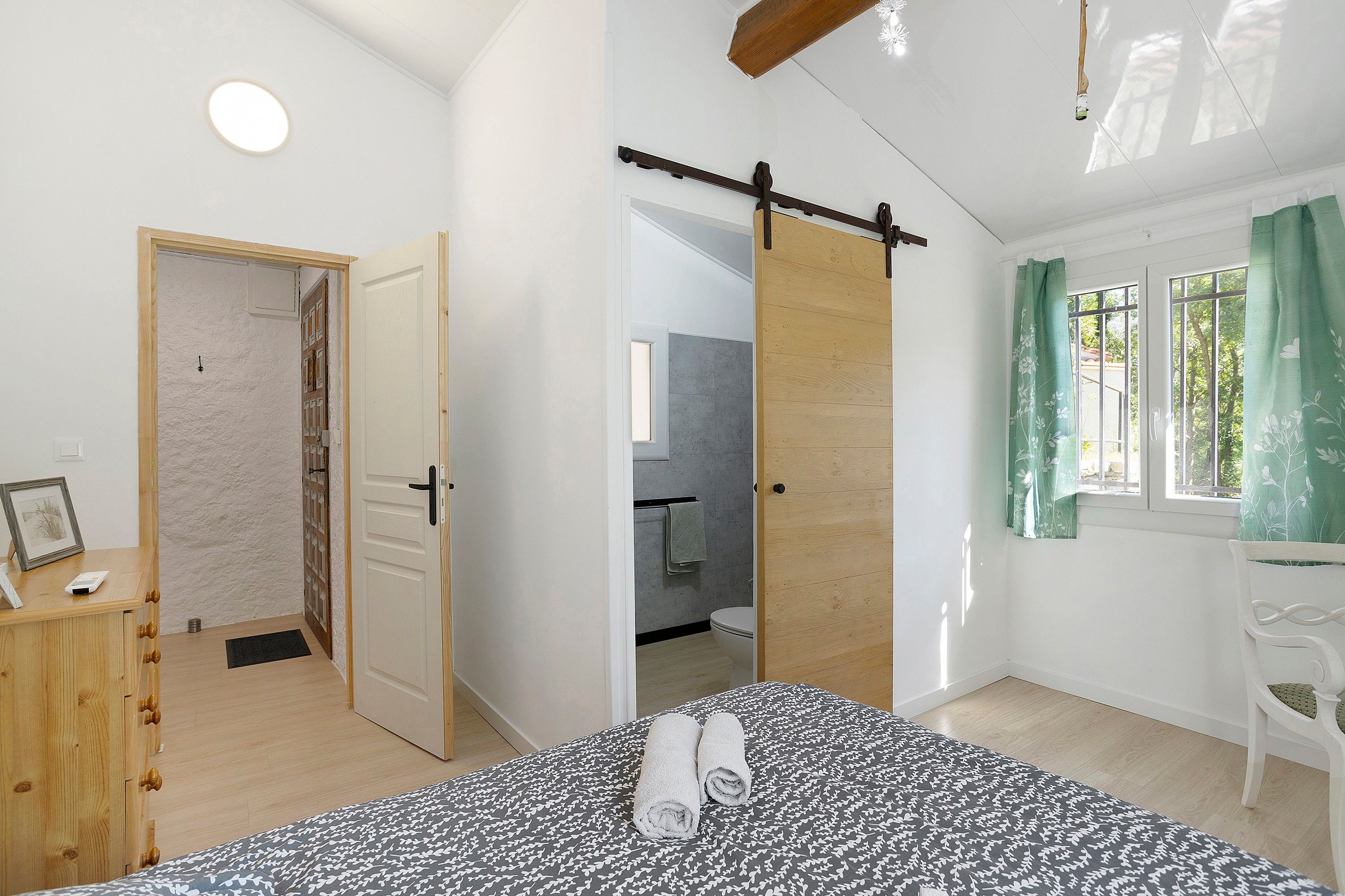 A bedroom with a bed, a wooden dresser, a bathroom with a toilet, and a window with curtains.