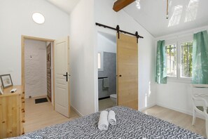 A bedroom with a bed, a wooden dresser, a bathroom with a toilet, and a window with curtains.