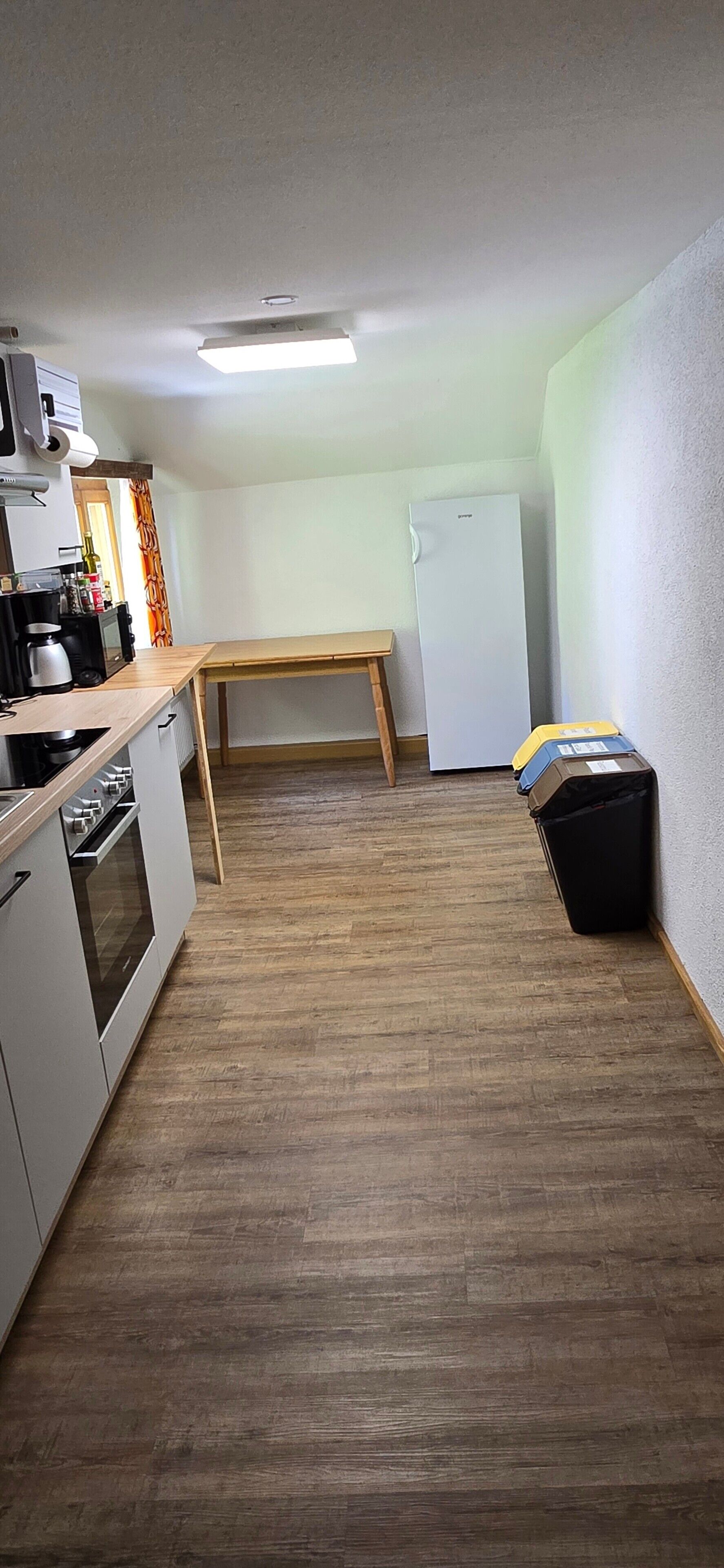 Una cocina compacta con una mesa de madera, armarios blancos, un refrigerador y un cubo de basura.