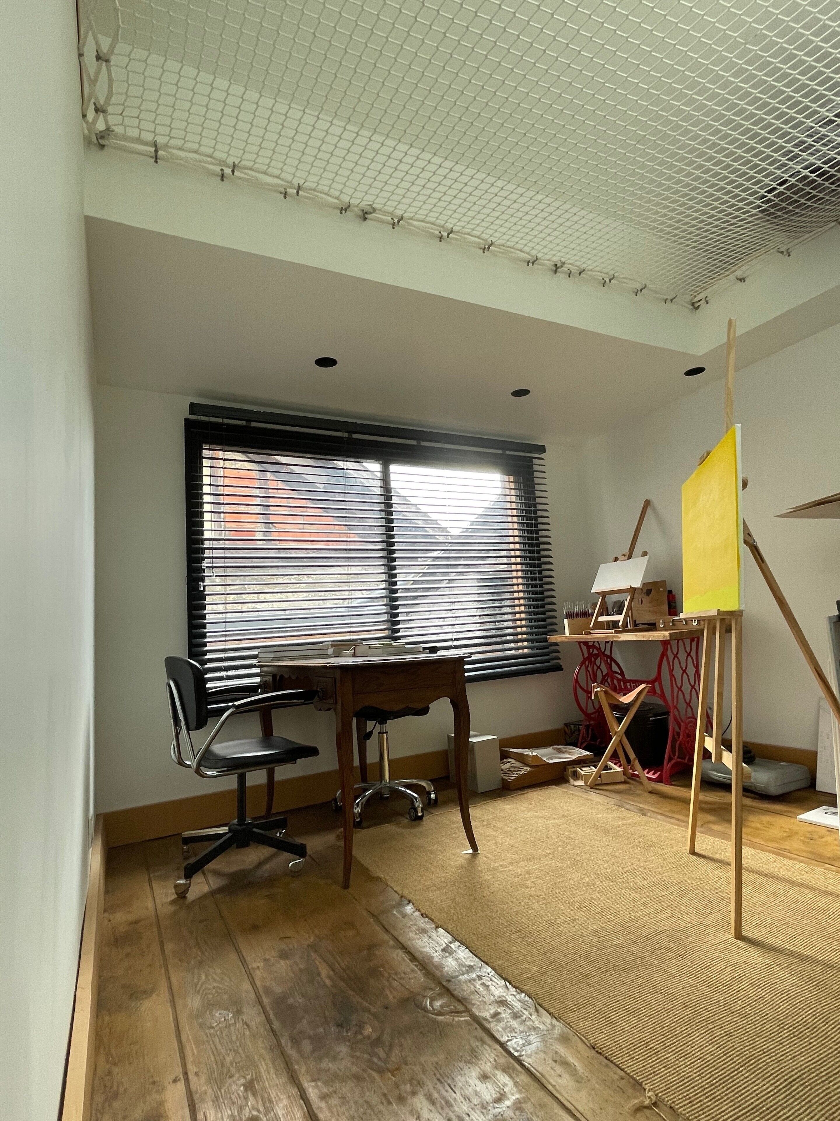 A room with a wooden desk, two chairs, a standing desk with a yellow board, and a window with blinds.