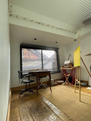 Um quarto com uma escrivaninha de madeira, duas cadeiras, uma mesa alta com um quadro amarelo e uma janela com persianas.