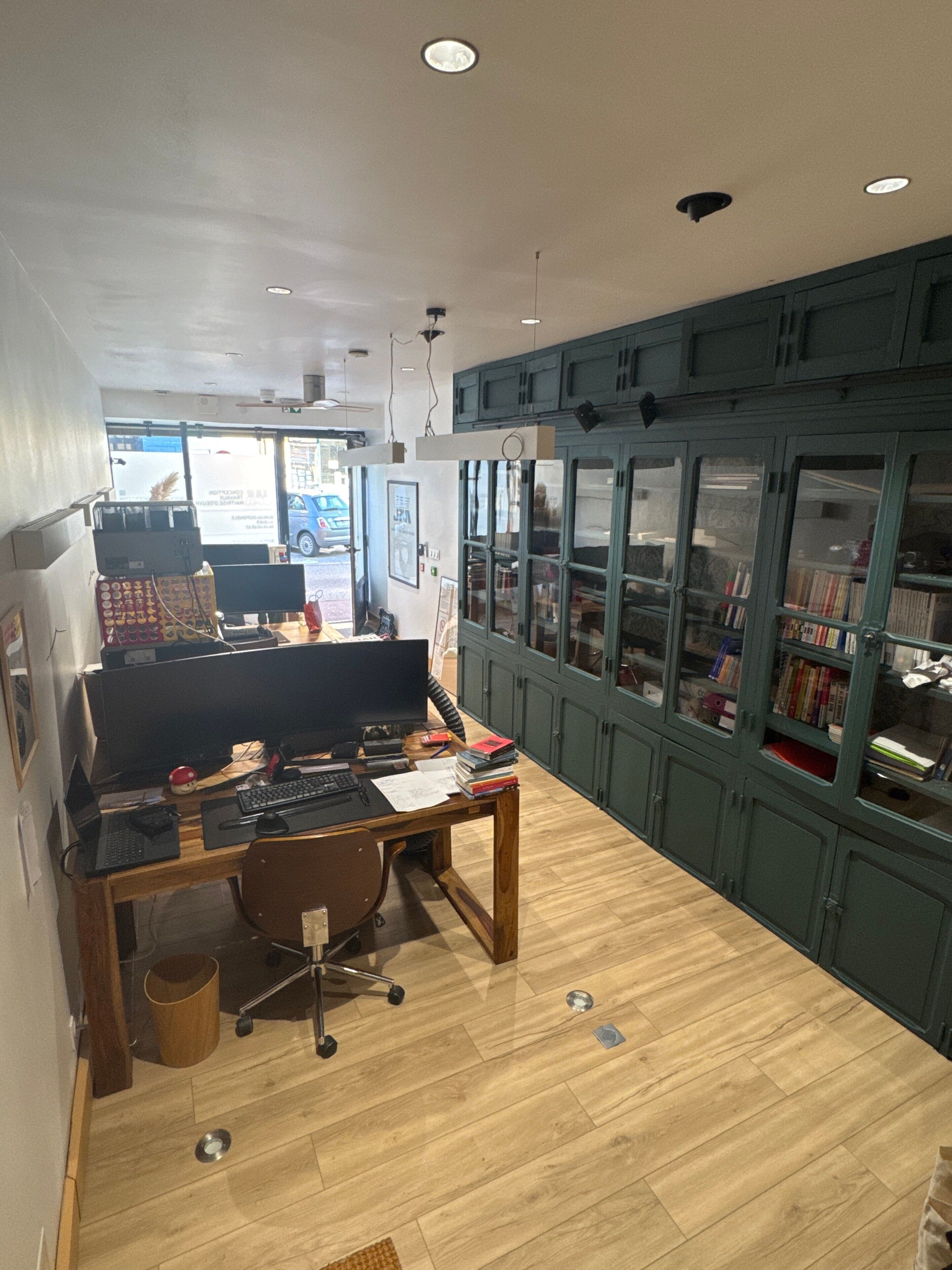 Une pièce avec un bureau en bois, un ordinateur et une chaise, une bibliothèque et une vue sur l’extérieur.