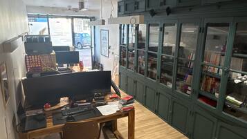Une pièce avec un bureau en bois, un ordinateur et une chaise, une bibliothèque et une vue sur l’extérieur.
