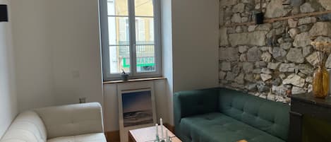 Una sala de estar con un sofá blanco, un sillón verde, una mesa de centro de madera, una pared de piedra y una ventana con vista a edificios.