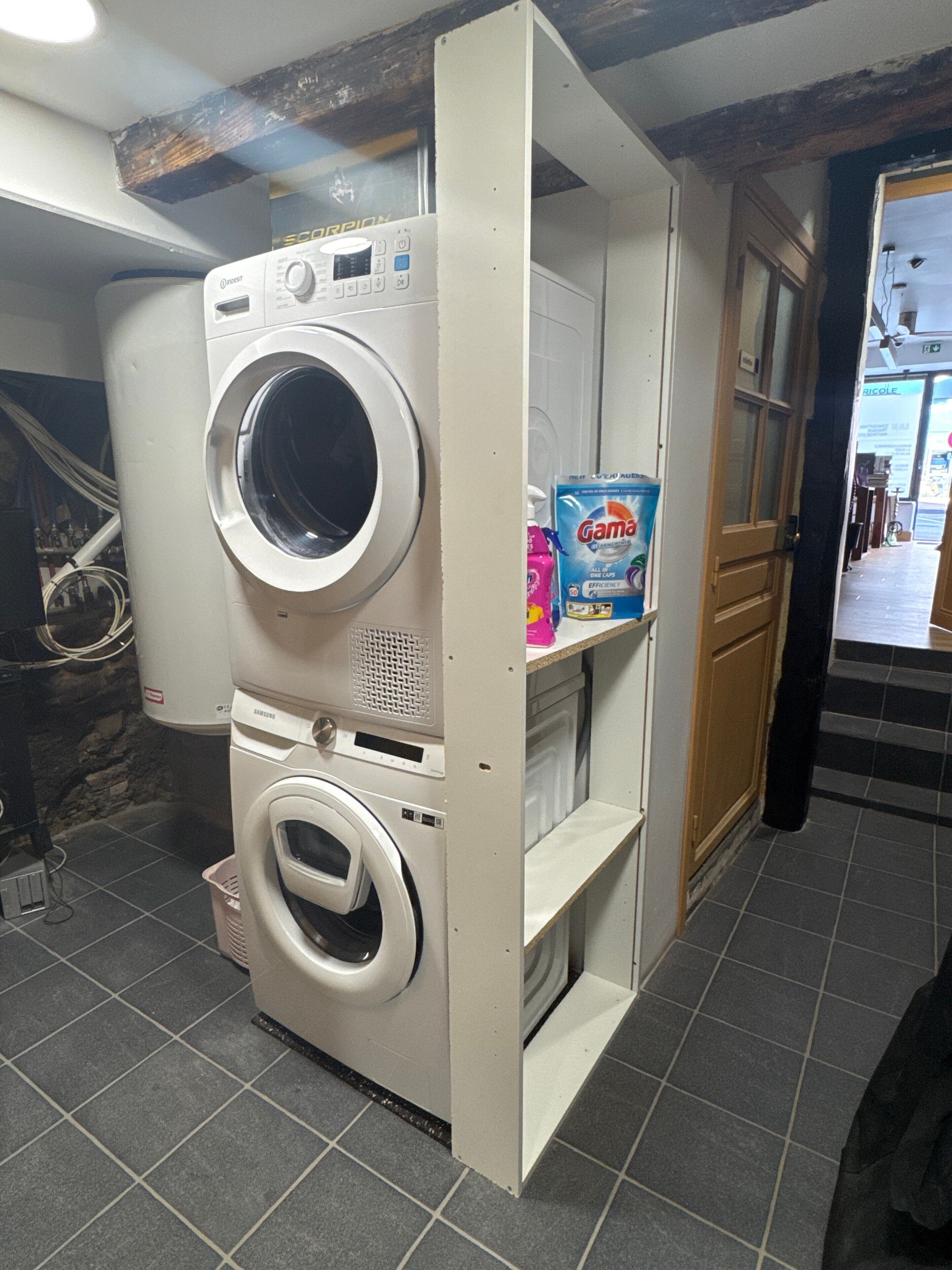 A laundry room with a washing machine and dryer, a shelf with detergent, and a door leading to another room.