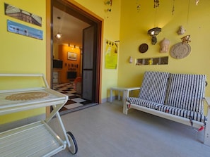 Sala de estar com sofá, mesinha e carrinho. Uma porta dá acesso a outro cômodo com piso xadrez.