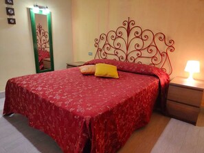 Quarto com colcha floral vermelha, cabeceira de metal, abajur e espelho na parede.