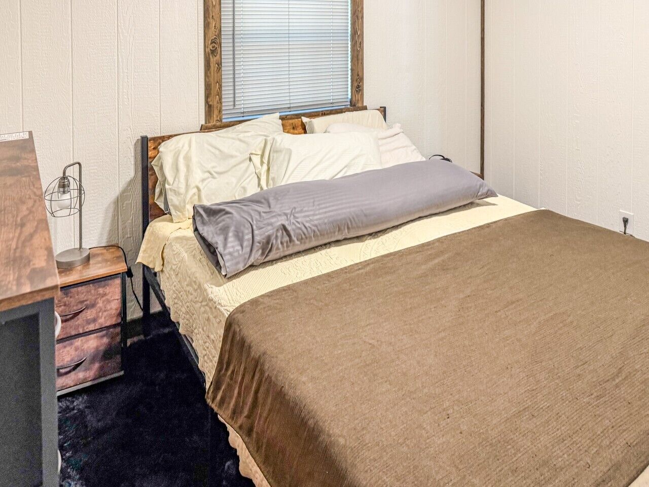 Ein Einzelbett mit brauner Bettdecke und grauem Kopfkissen, ein Nachttisch aus Holz mit einer Lampe und ein Fenster mit Jalousien.