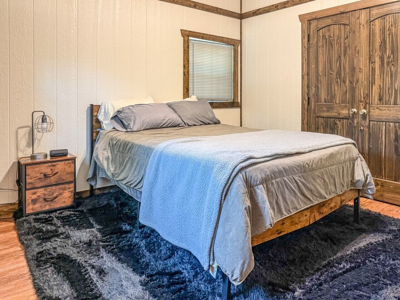 Een slaapkamer met een bed, nachtkastje, een houten garderobe en een raam met jaloezieën.