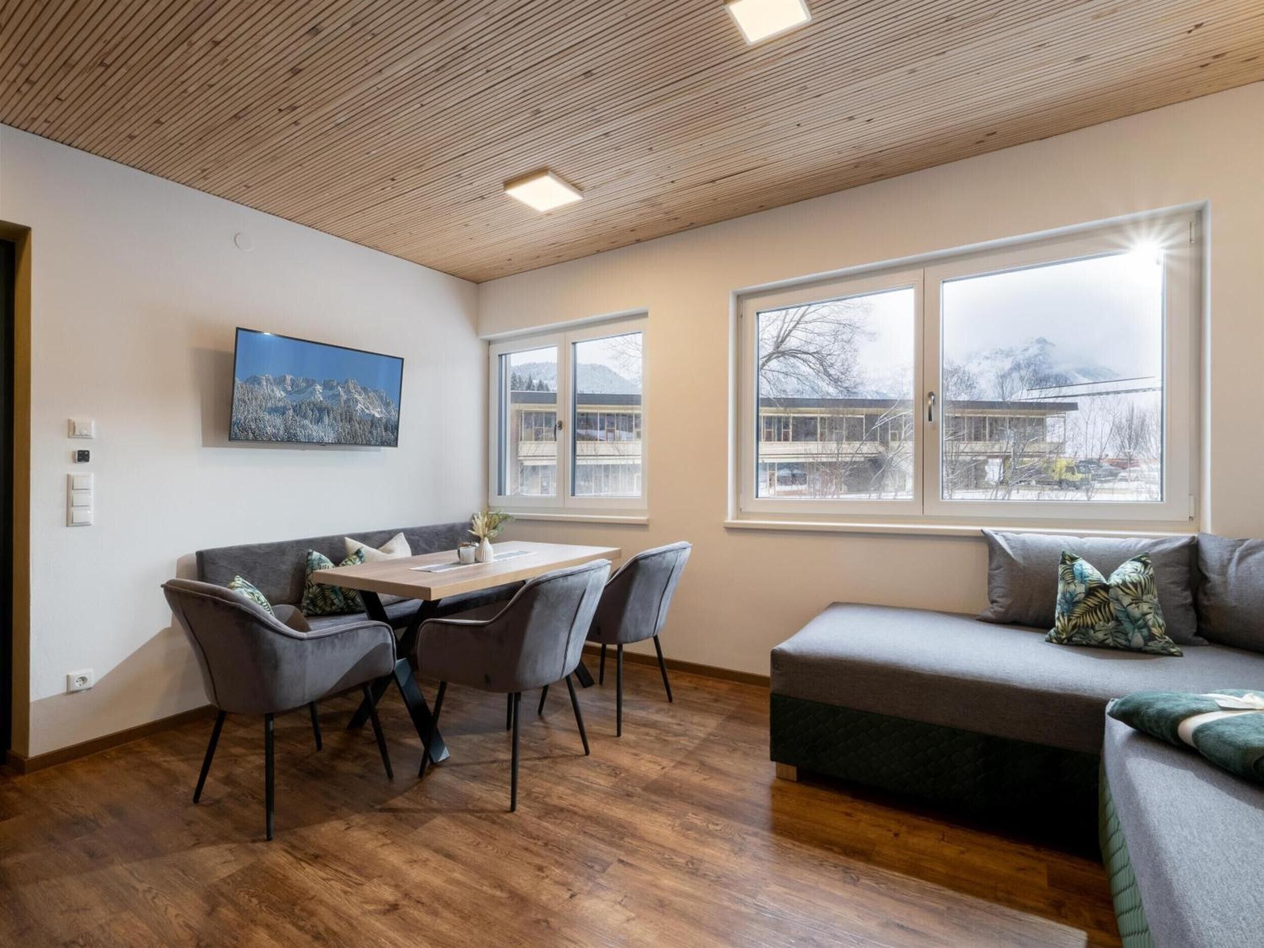 Un soggiorno moderno con soffitto in legno, un tavolo da pranzo con sedie, un divano e ampie finestre con vista su edifici e montagne.