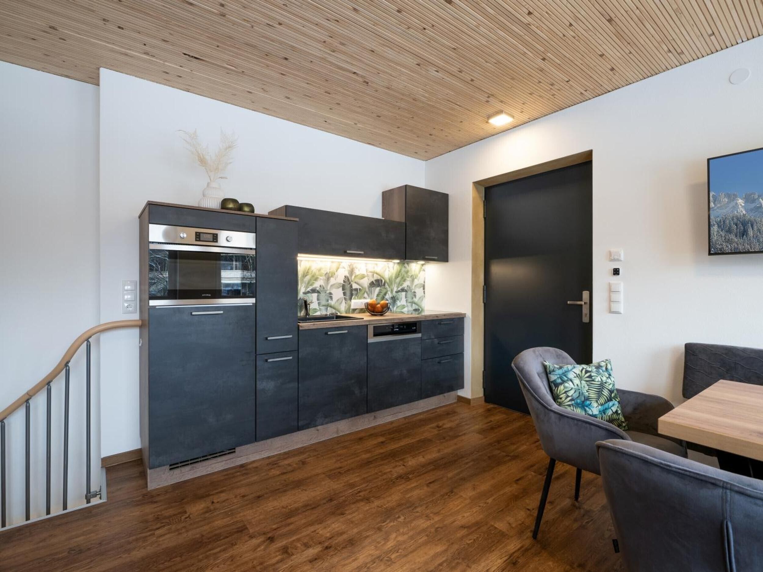 Una cucina moderna con mobili scuri, forno integrato e lavastoviglie. Una scala in legno conduce a un altro livello.