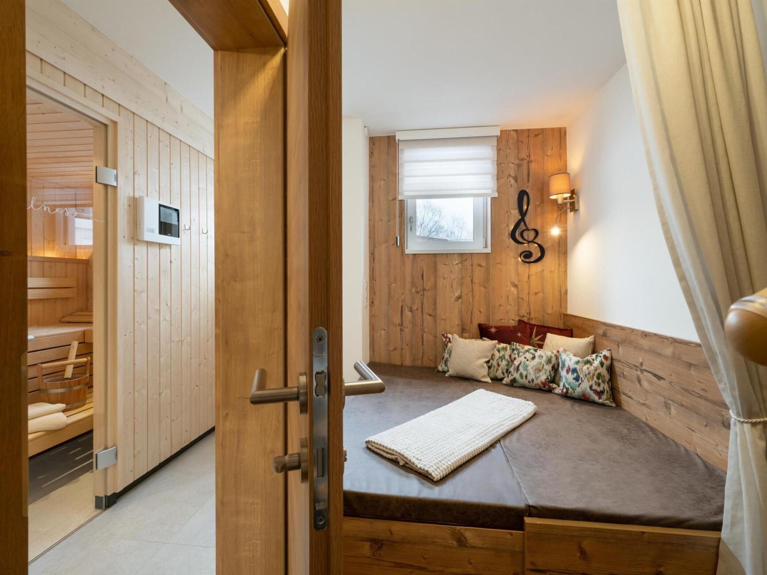 Ein Zimmer mit einer hölzernen Sitzbank, einem Bett mit brauner Matratze, einem Fenster mit Jalousien und einer an der Wand befestigten Leuchte.