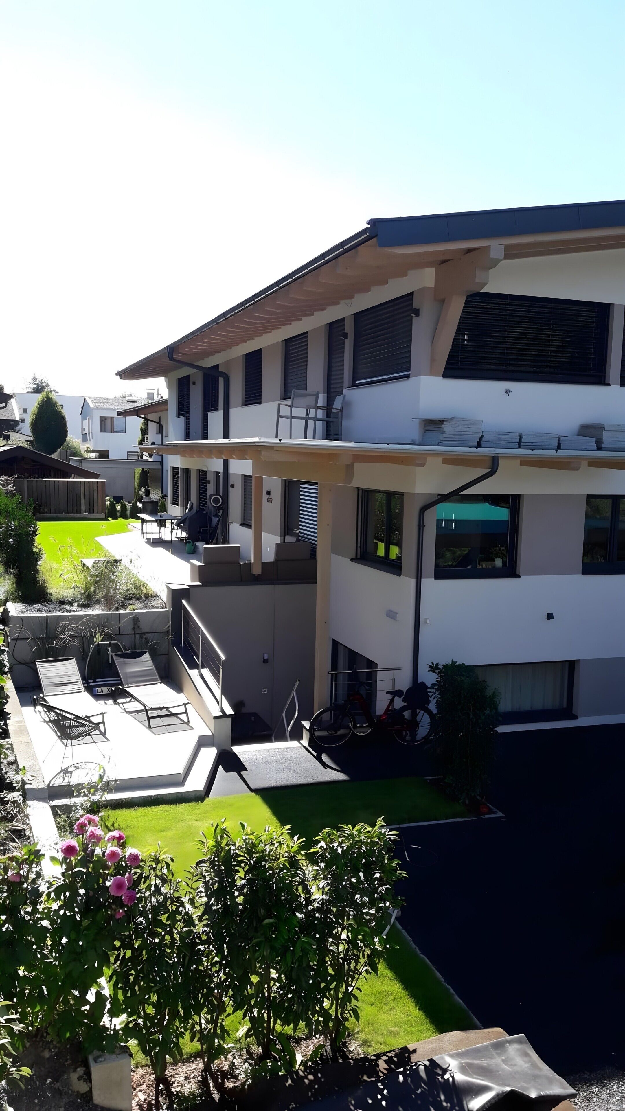 Un edificio moderno a due piani con tetto piatto, ampie finestre e un’area salotto esterna coperta con sedie e tavoli.