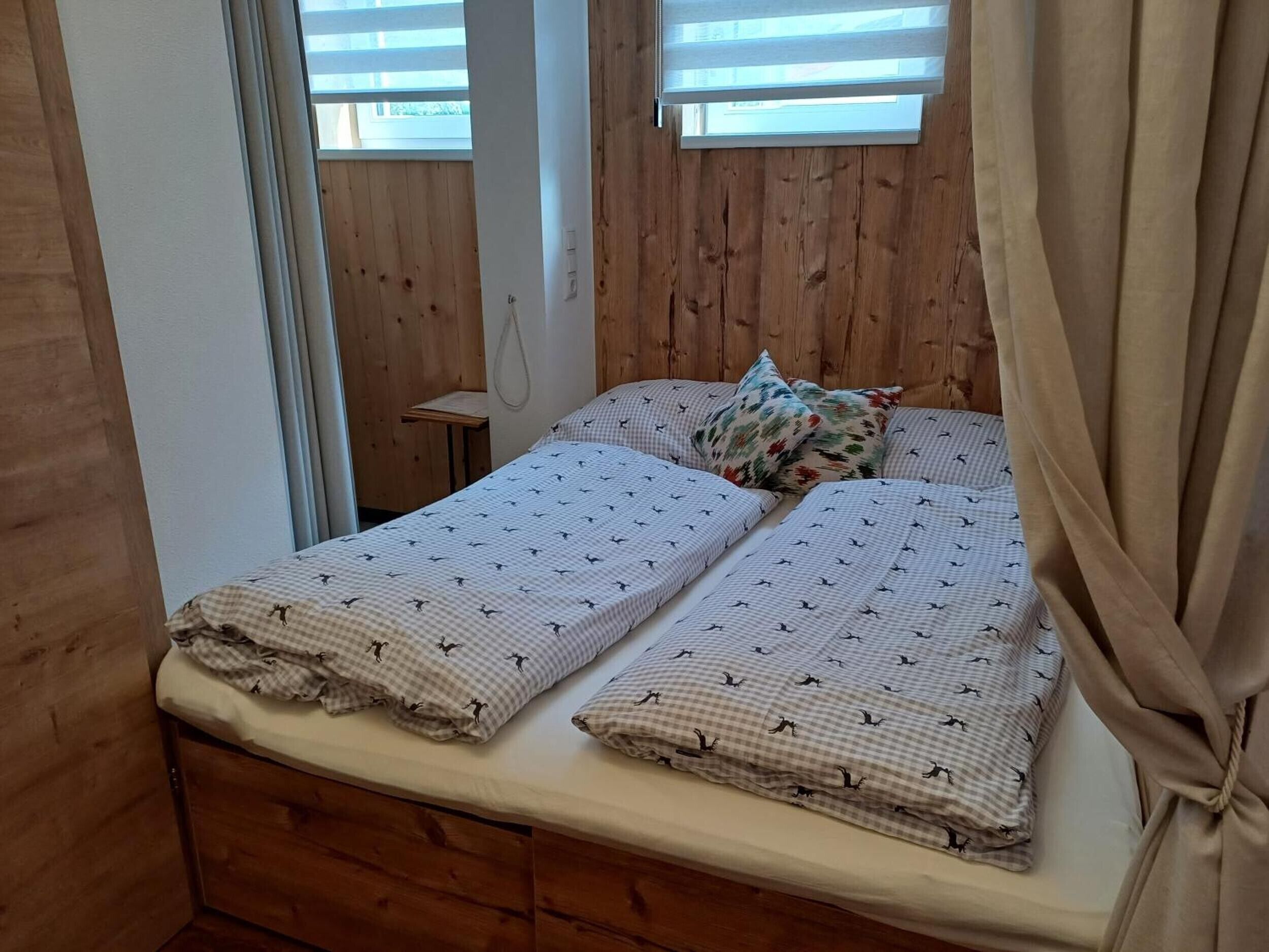 Un lit simple avec une couvre-lit à carreaux et des coussins assortis, une tête de lit en bois et une fenêtre avec des stores.