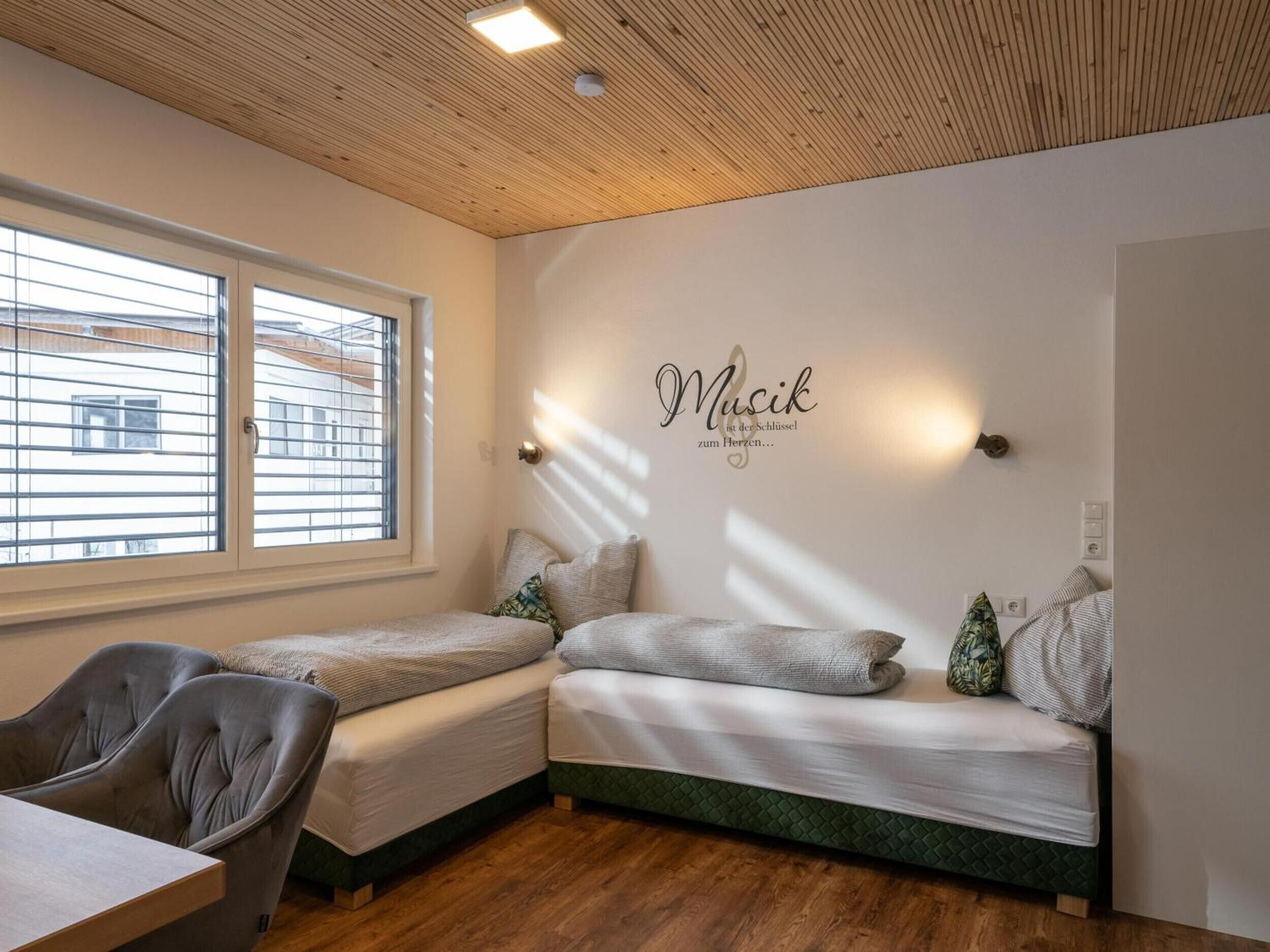 A bedroom with two single beds, a wooden ceiling, a wall-mounted light, and a window with blinds.