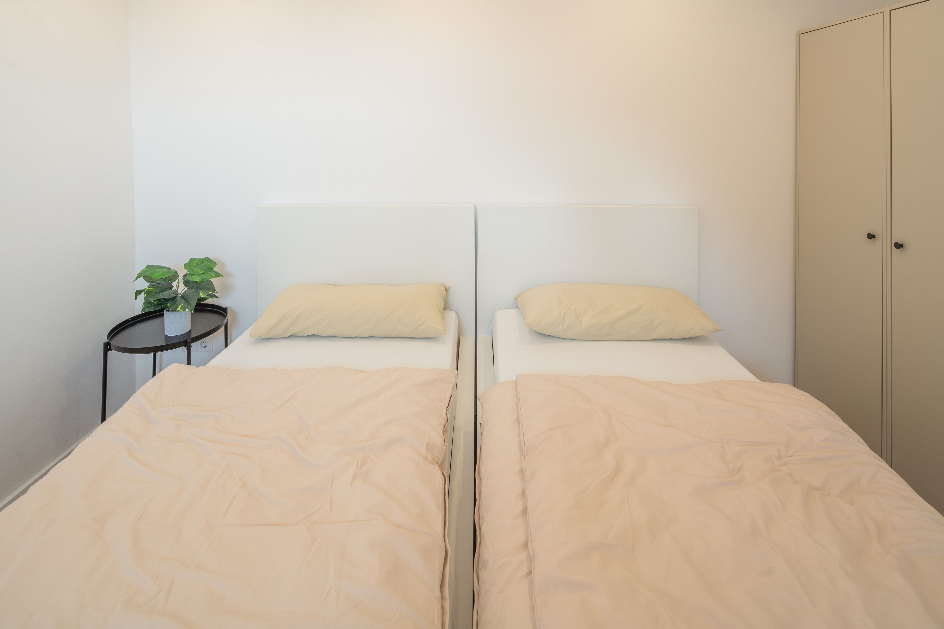 Un letto matrimoniale con copripiumino e cuscini color beige, un tavolino rotondo con una pianta in vaso e un armadio con maniglia.