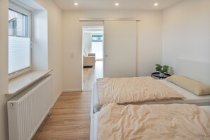 Un dormitori modern amb dos llits, un radiador, una finestra amb persianes i vista a una altra habitació.