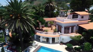Una casa de dos pisos con piscina, rodeada de palmeras y otra vegetación.