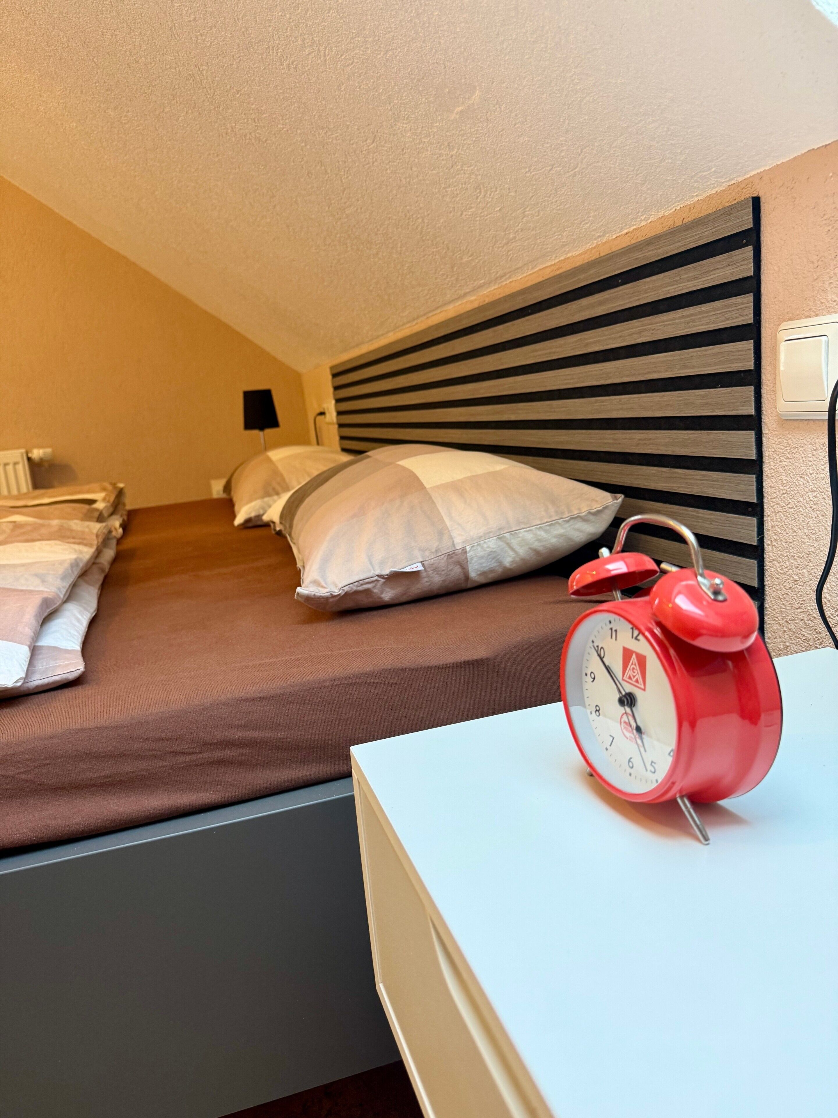 A hotel room with a bed, pillows, a bedside table, and a red alarm clock.