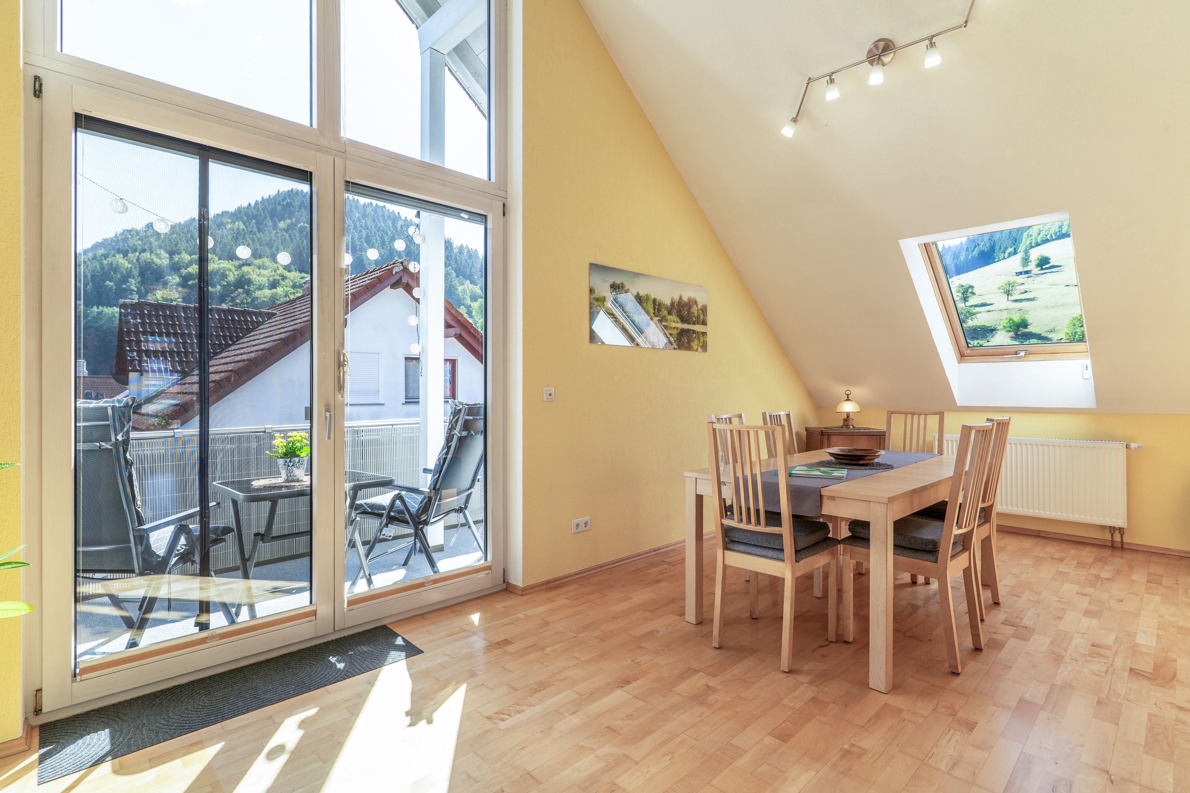 A dining area with a table and chairs, a window with a view of a house and trees, and a ceiling light fixture.