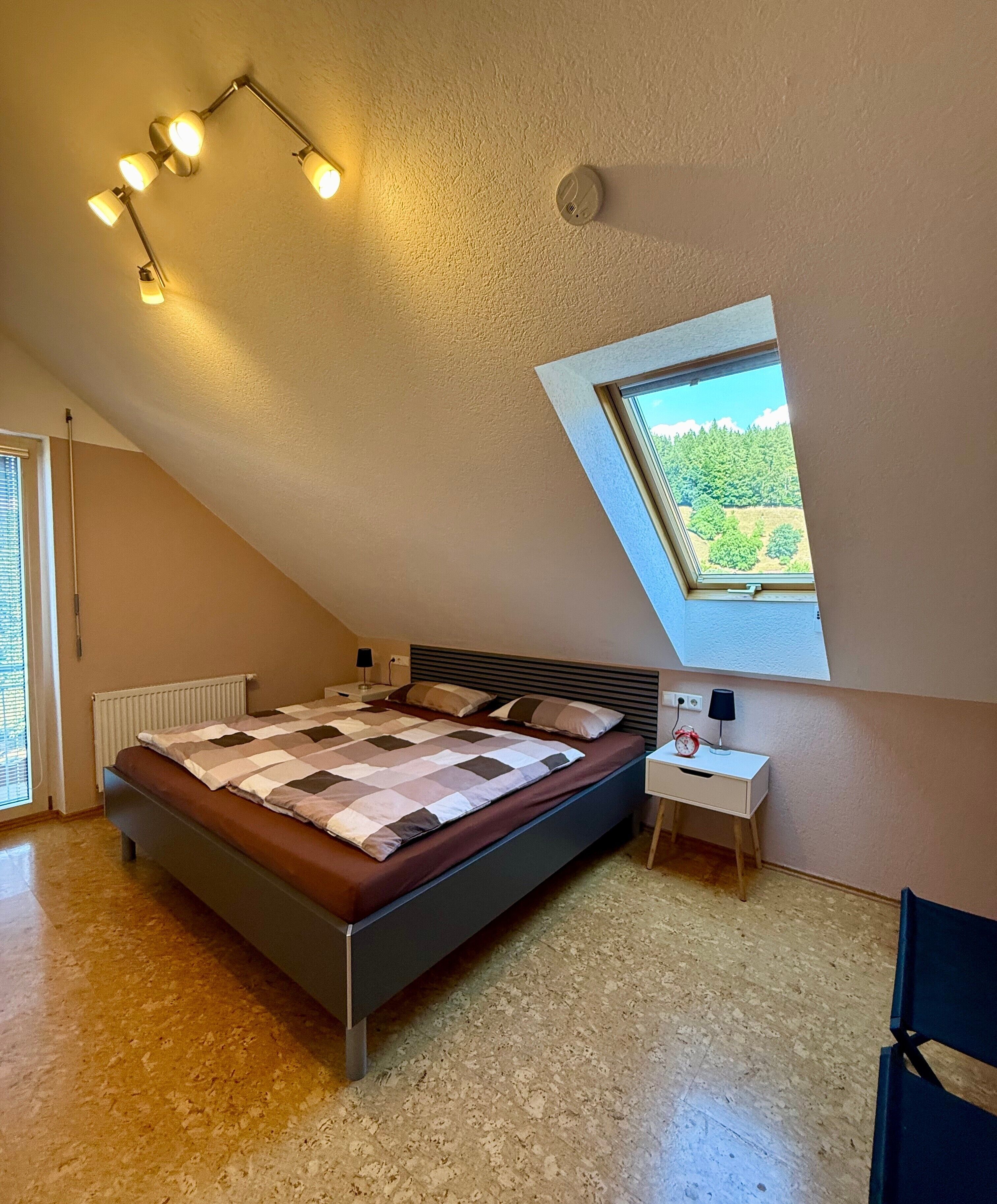 A bedroom with a sloped ceiling, a skylight, a bed with a quilt, a bedside table with a lamp, and a chair.