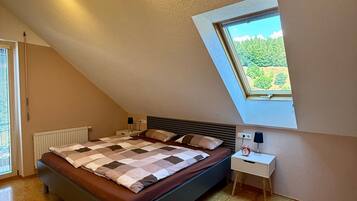 A bedroom with a sloped ceiling, a skylight, a bed with a quilt, a bedside table with a lamp, and a chair.