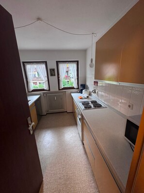 A compact kitchen with a built-in stove, a radiator, and two windows with curtains.