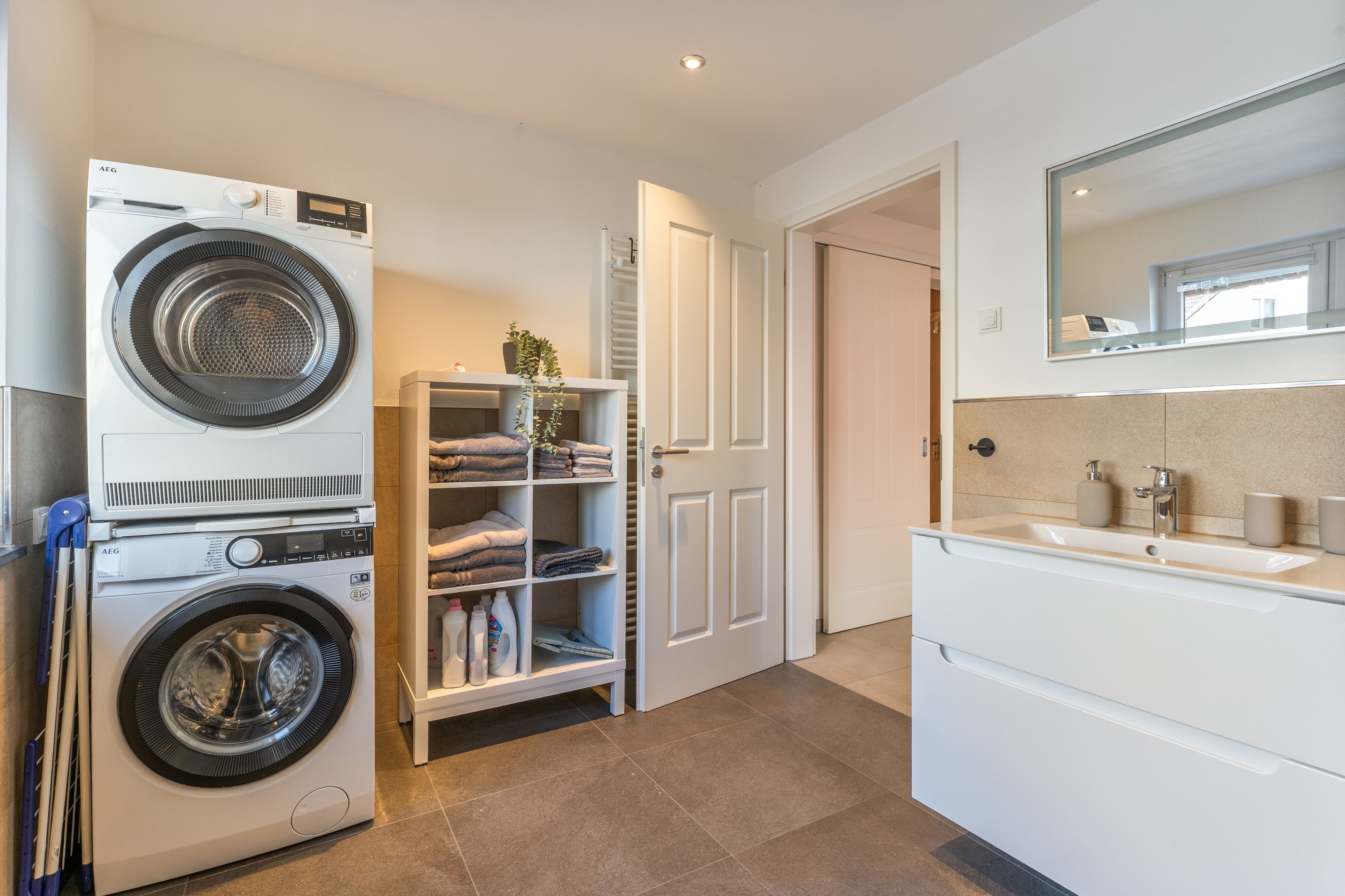 A laundry room with a front-loading washing machine, a drying rack, and a shelf with folded towels.