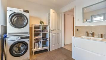 A laundry room with a front-loading washing machine, a drying rack, and a shelf with folded towels.