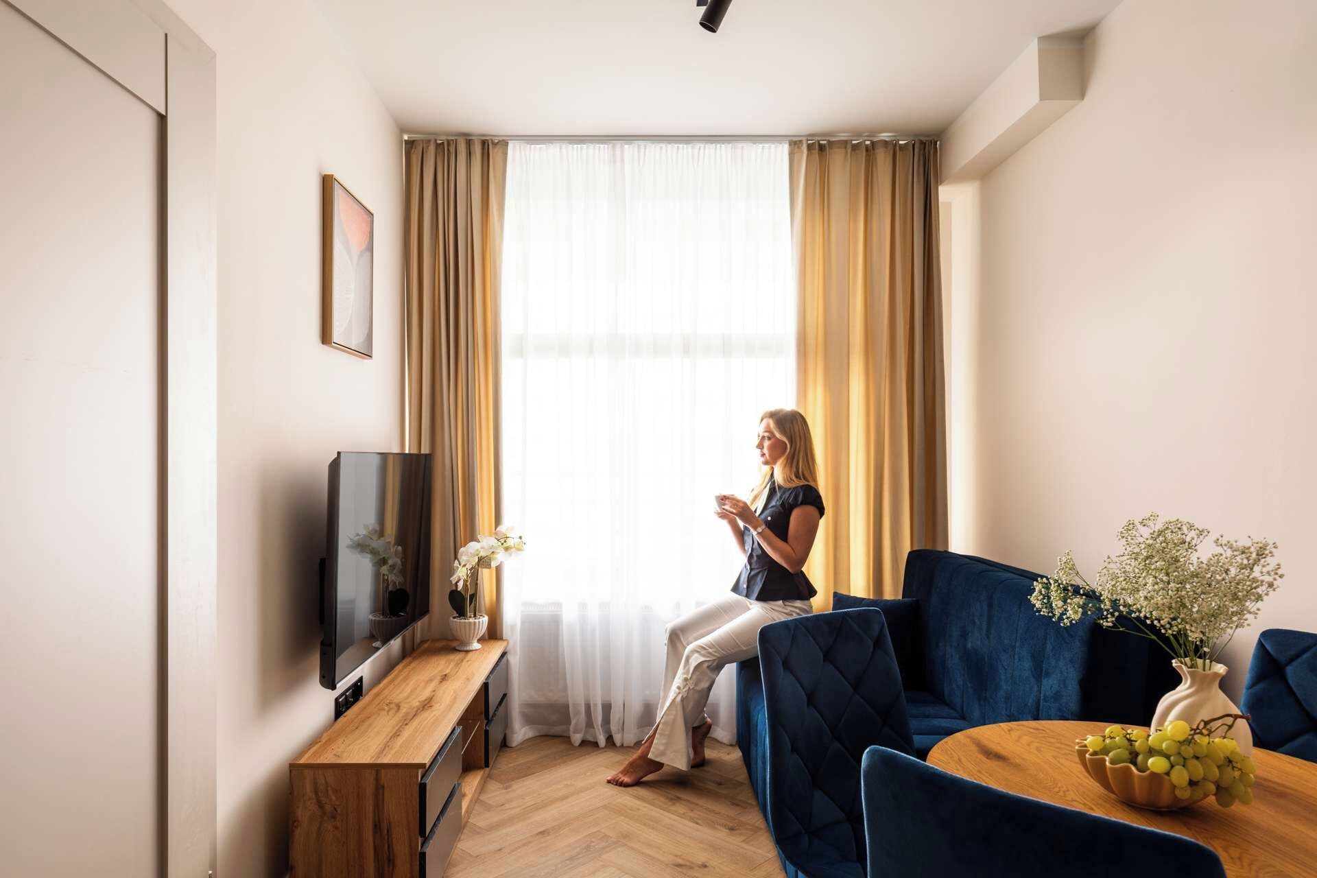 A modern living room with a sofa, a coffee table with a vase of flowers, a television, and a window with curtains.