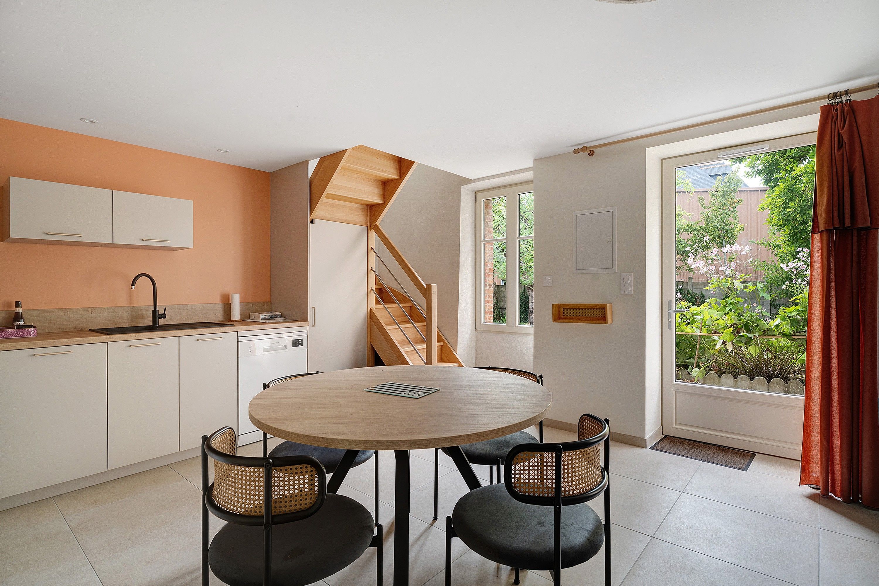 Une cuisine moderne avec une table de salle à manger ronde, deux chaises, un escalier et une vue sur un jardin, visible à travers une grande fenêtre.