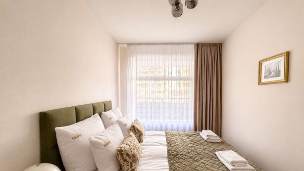 Una camera d'albergo con un letto, una lampada da comodino, una finestra con tende e un quadro appeso al muro.