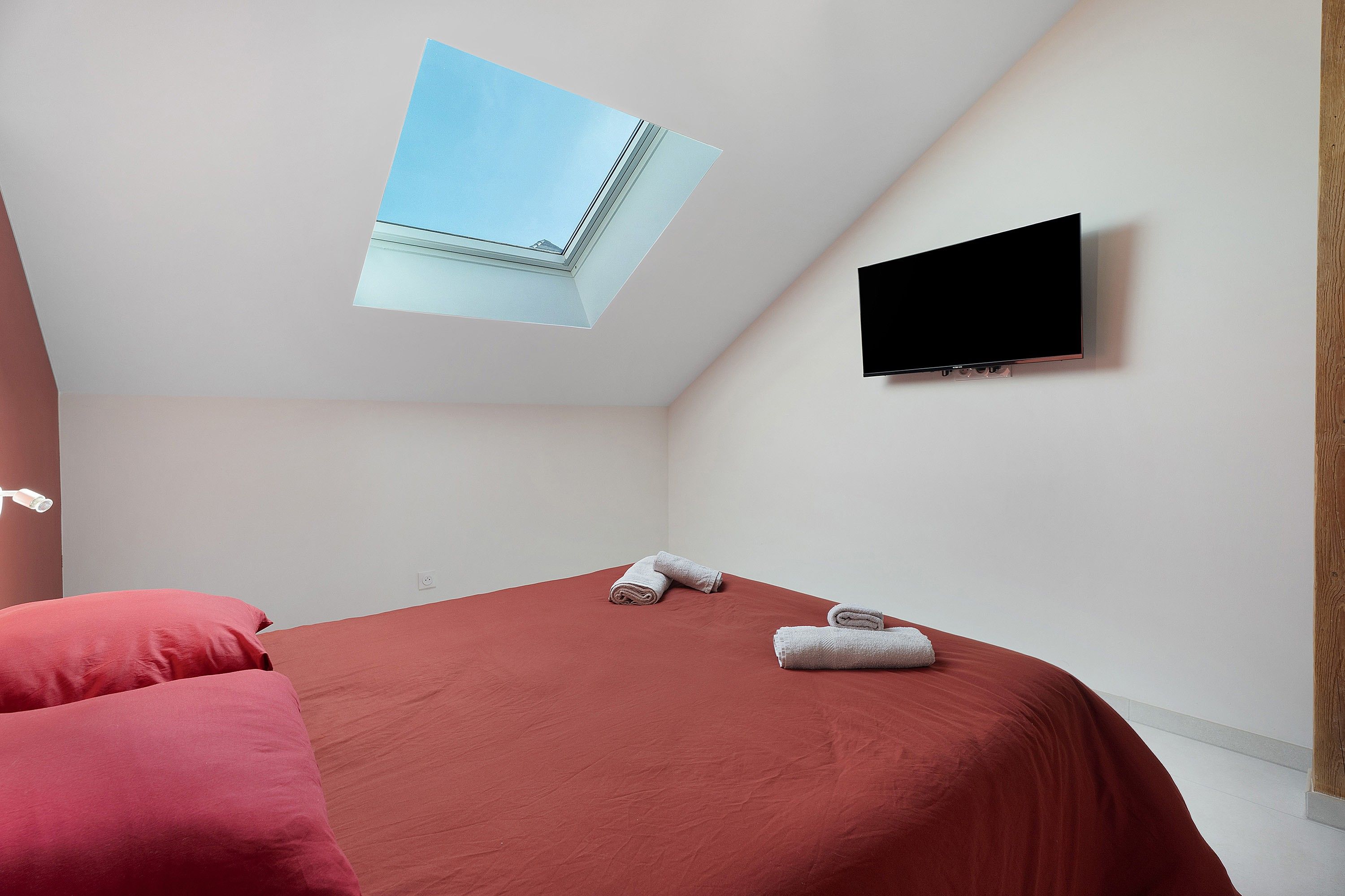 A room with a bed, a flat-screen TV mounted on the wall, and a skylight.
