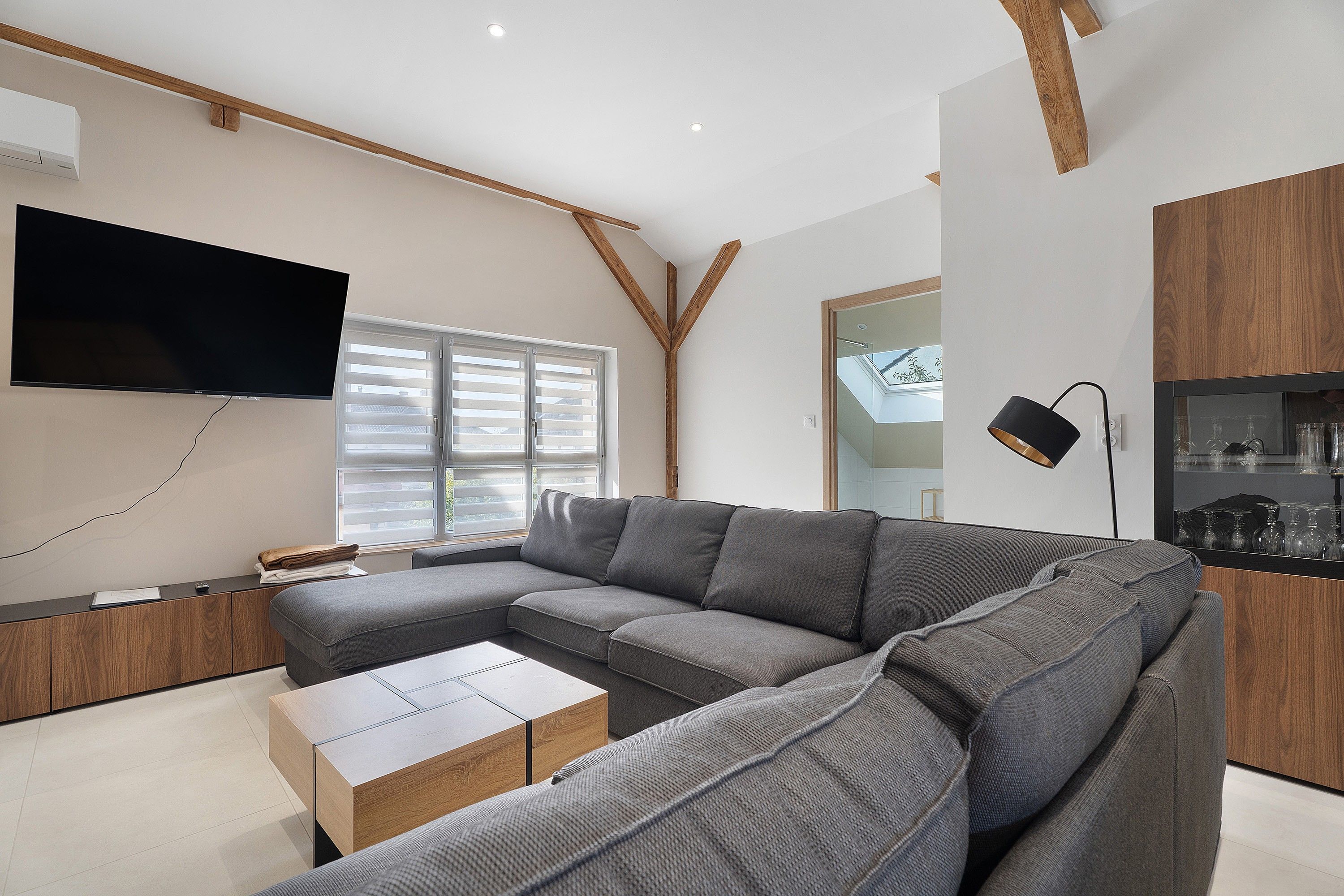 Un salon moderne avec un canapé d’angle, un téléviseur à écran plat fixé au mur et un meuble en bois doté d’une porte en verre.
